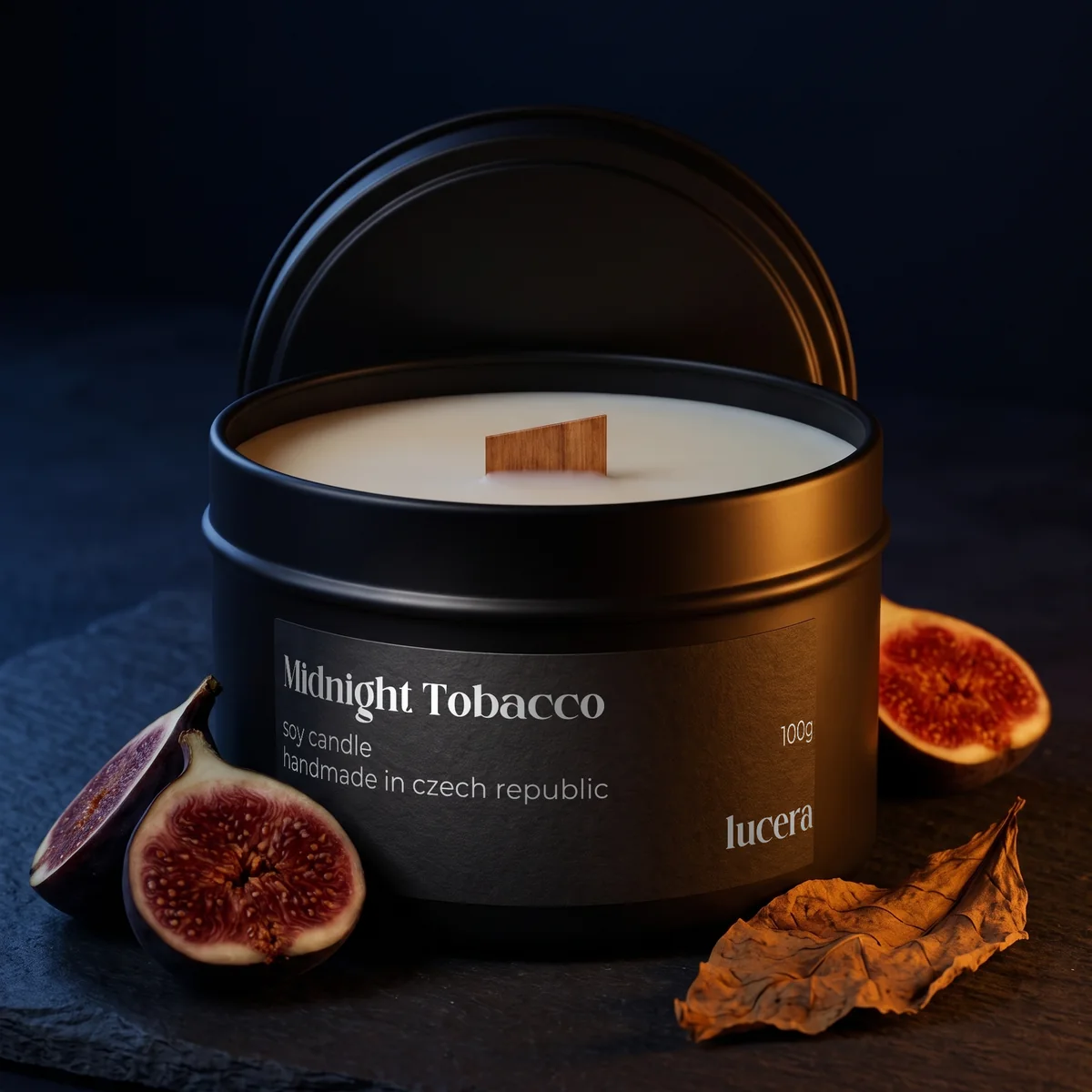 Svíčka Lucera Midnight Tobacco v černé plechovce na tmavě modré břidlici, vedle rozkrojený fík a sušený tabákový list