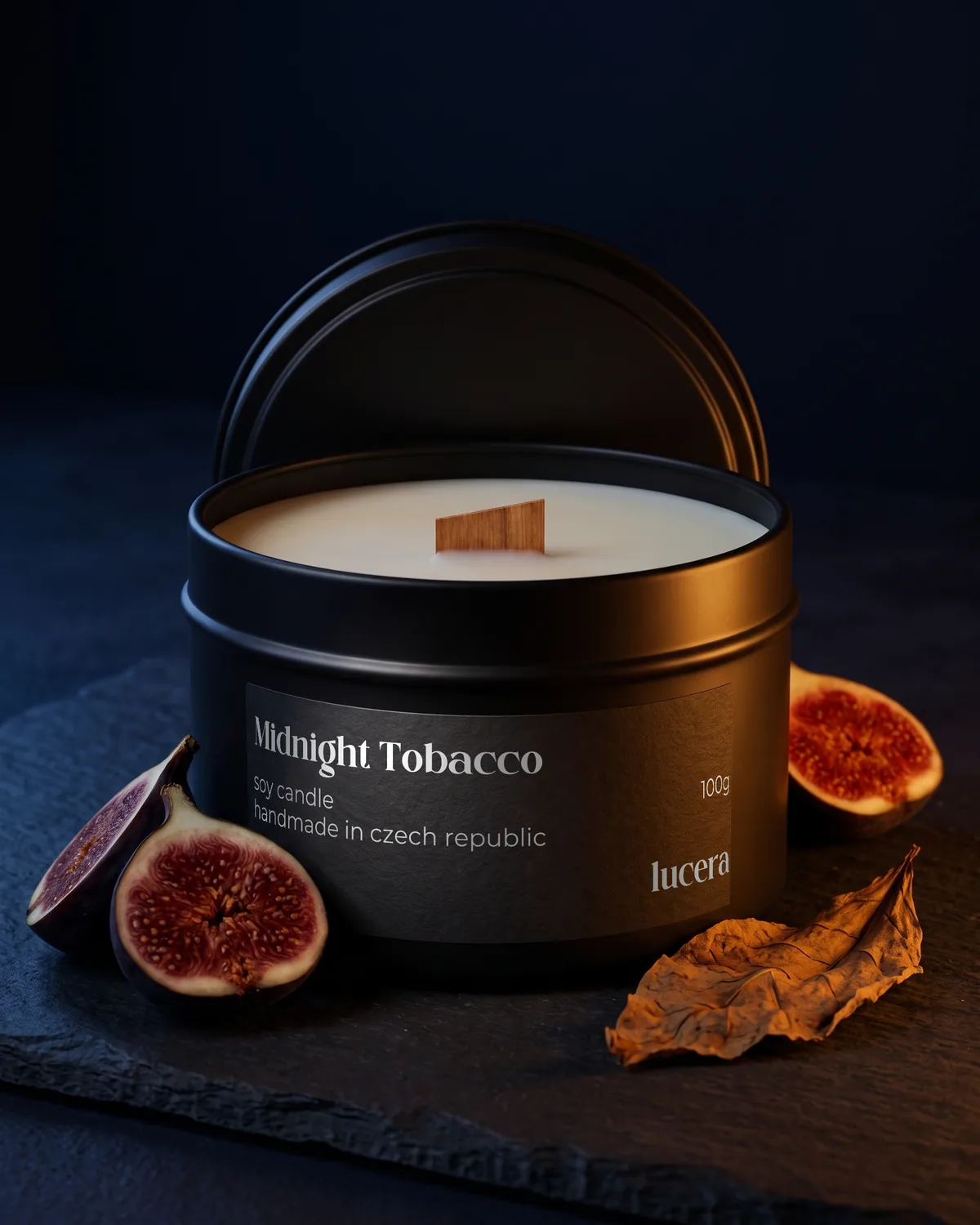 Svíčka Lucera Midnight Tobacco v černé plechovce na tmavě modré břidlici, vedle rozkrojený fík a sušený tabákový list