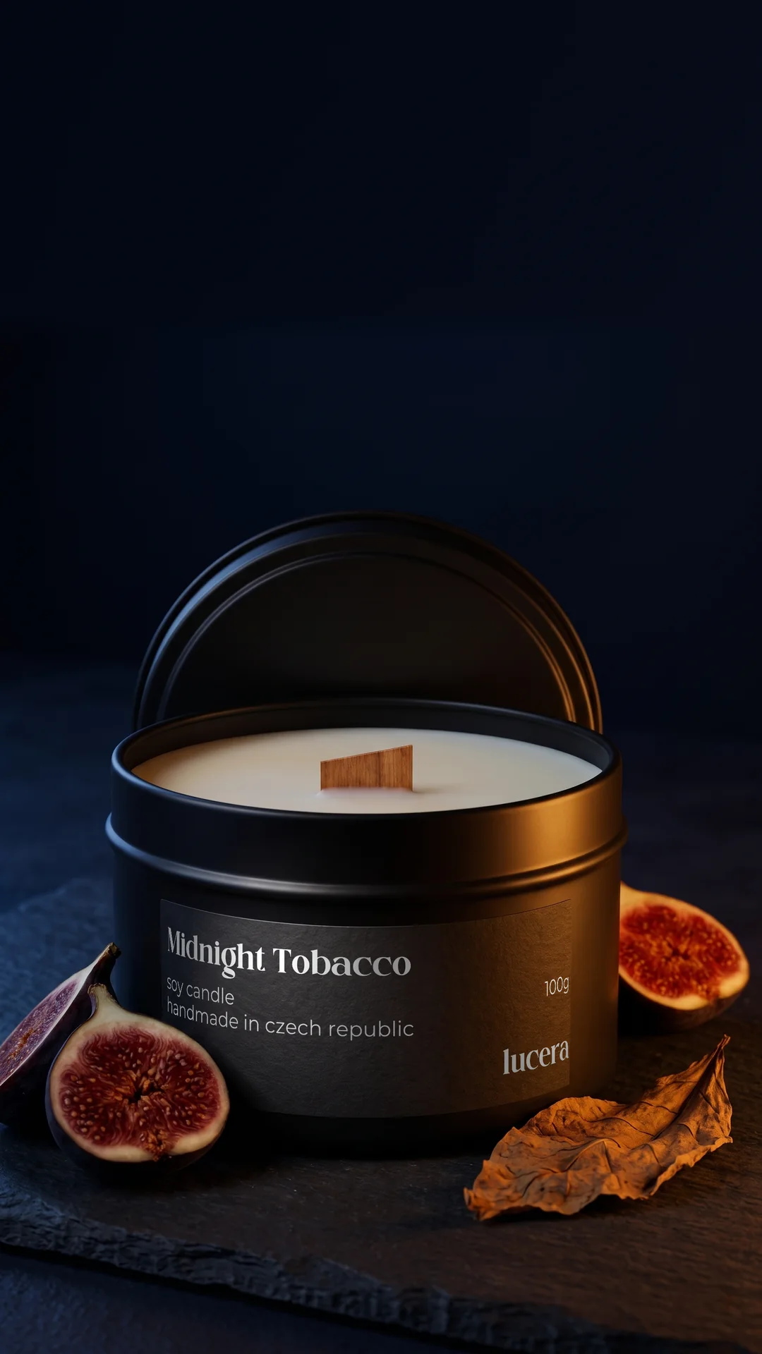 Svíčka Lucera Midnight Tobacco v černé plechovce na tmavě modré břidlici, vedle rozkrojený fík a sušený tabákový list