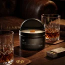 Svíčka Lucera Noir v černé plechovce na dubovém stole vedle sklenice whisky a zápalek, v pozadí kožené křeslo