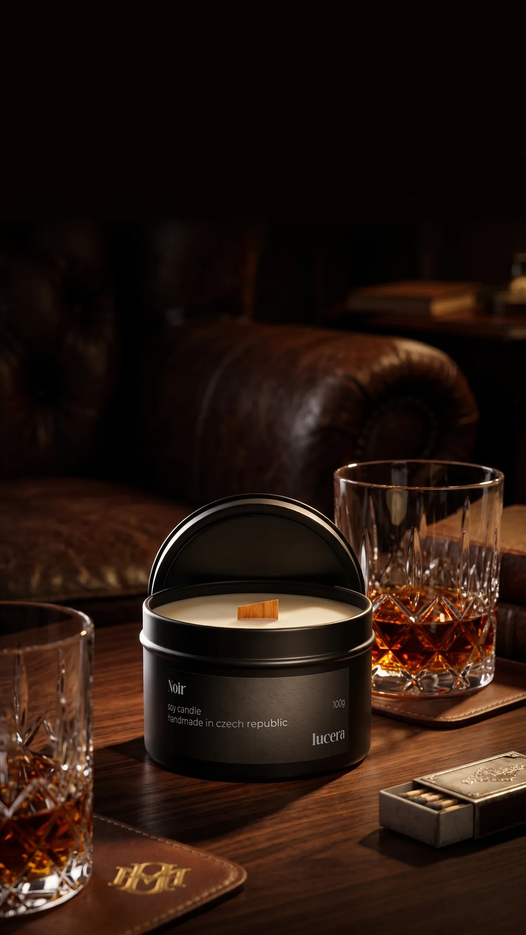 Svíčka Lucera Noir v černé plechovce na dubovém stole vedle sklenice whisky a zápalek, v pozadí kožené křeslo