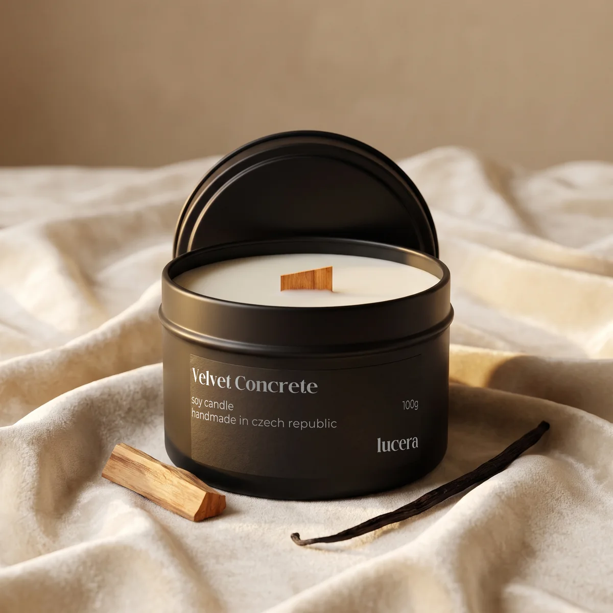 Svíčka Lucera Velvet Concrete v černé plechovce na krémové látce vedle dřívka palo santo a vanilkového lusku