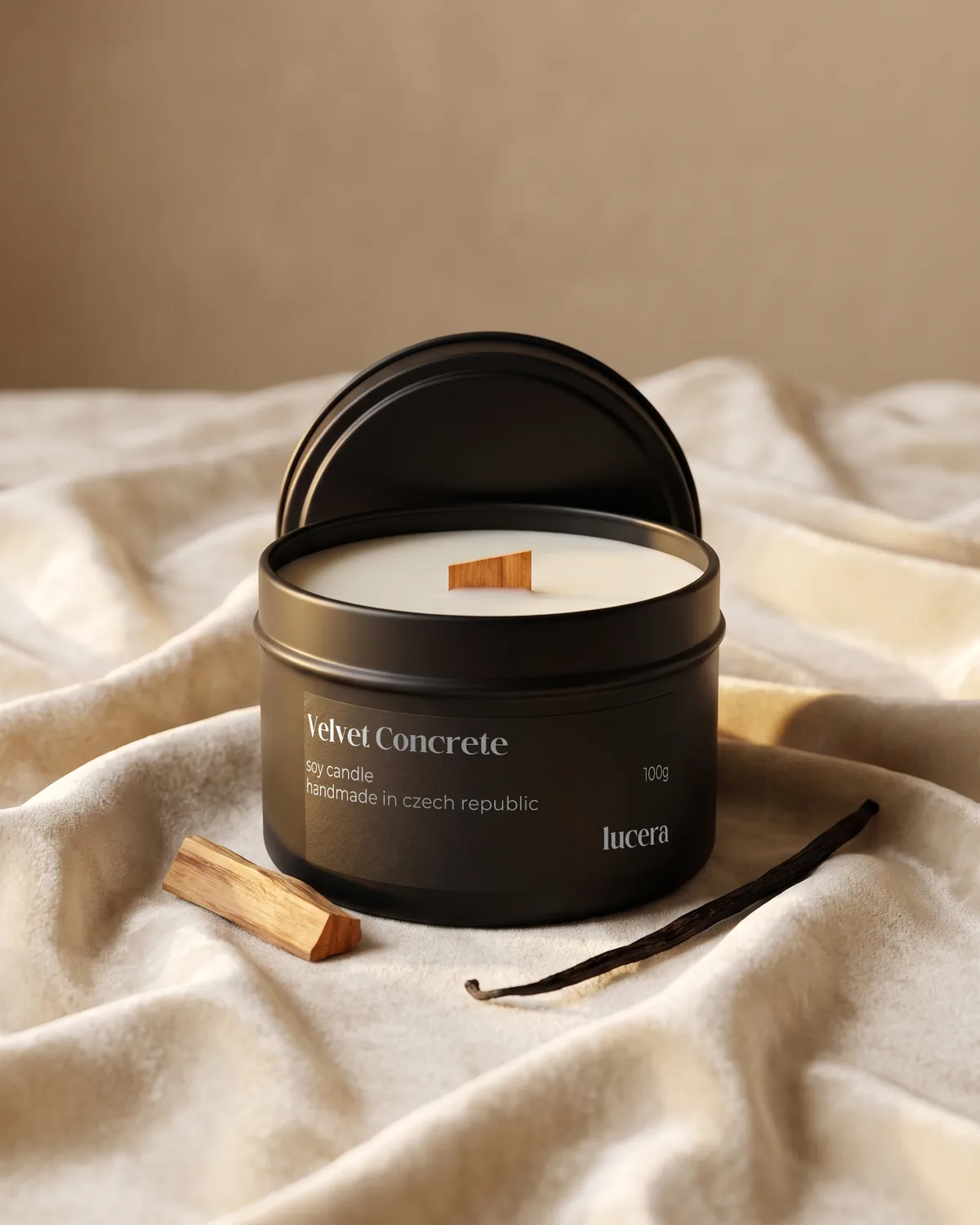 Svíčka Lucera Velvet Concrete v černé plechovce na krémové látce vedle dřívka palo santo a vanilkového lusku